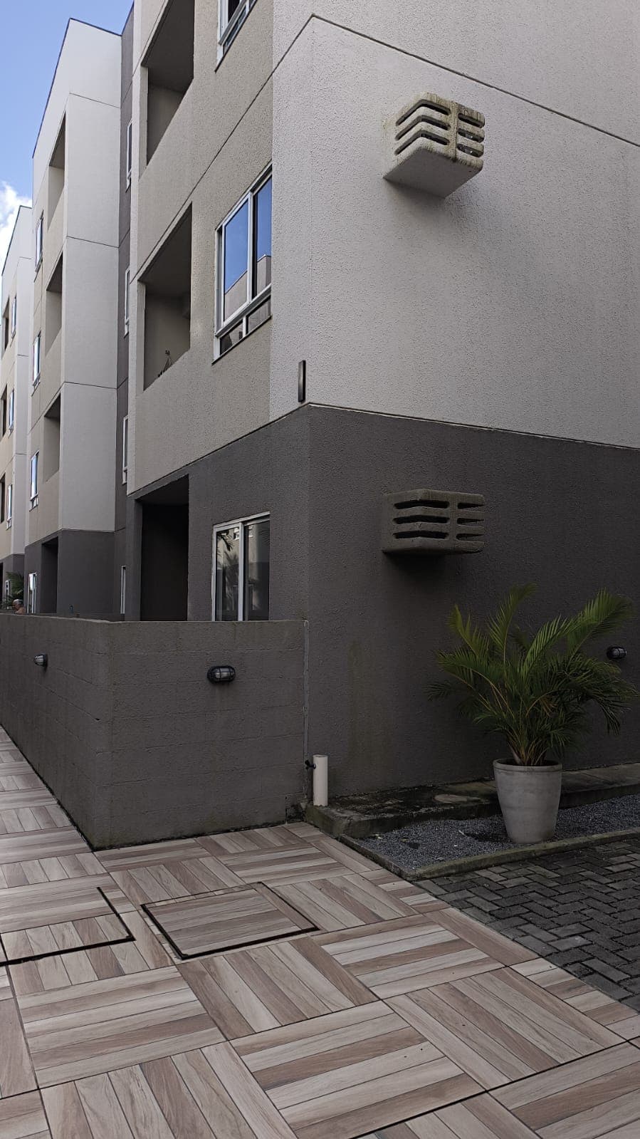 Fachada externa do edifício com piso revestido e paisagismo em vaso.